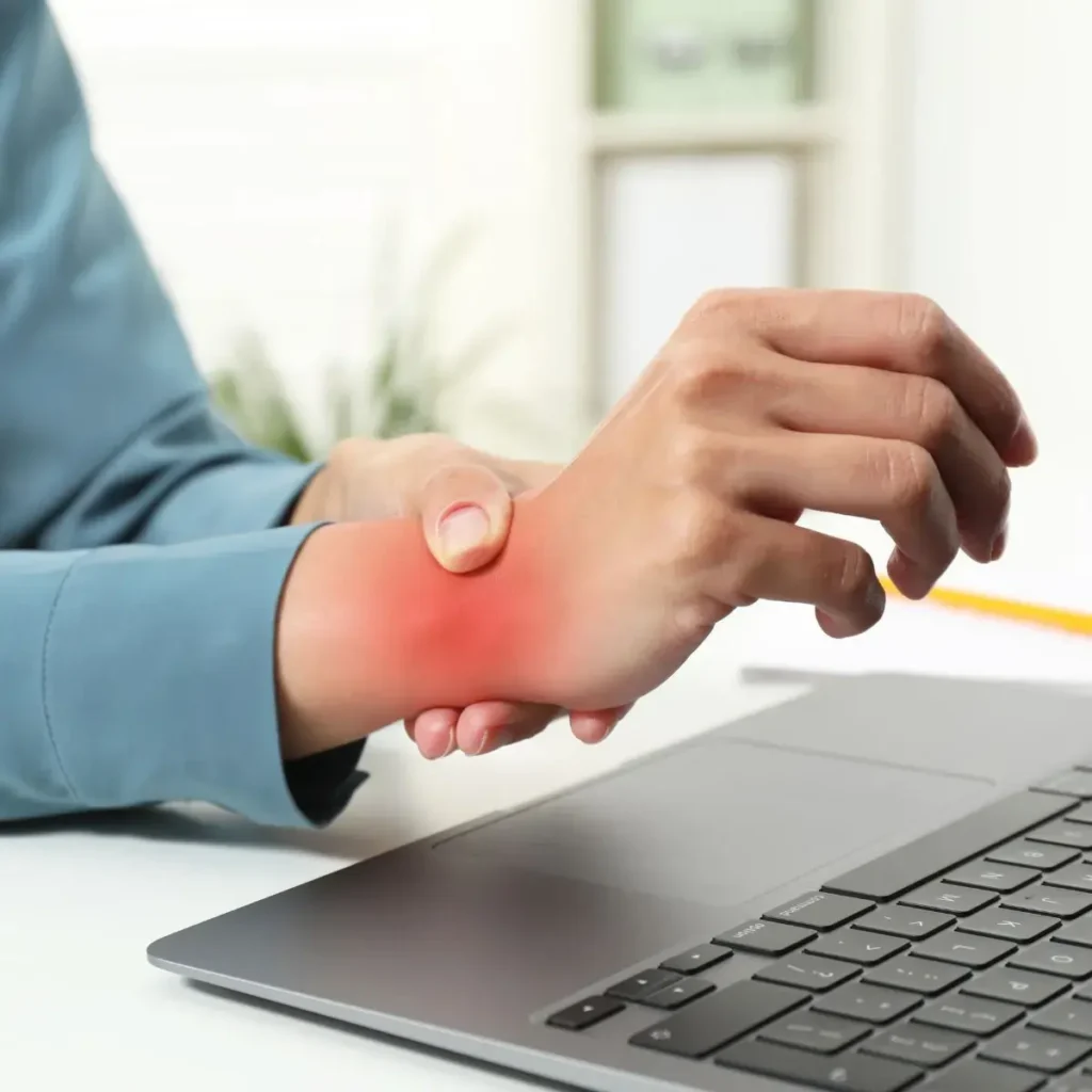 A person holds their painful wrist with one hand, showing redness around the joint, while sitting at a desk with a laptop, suggesting wrist pain or injury possibly from typing.