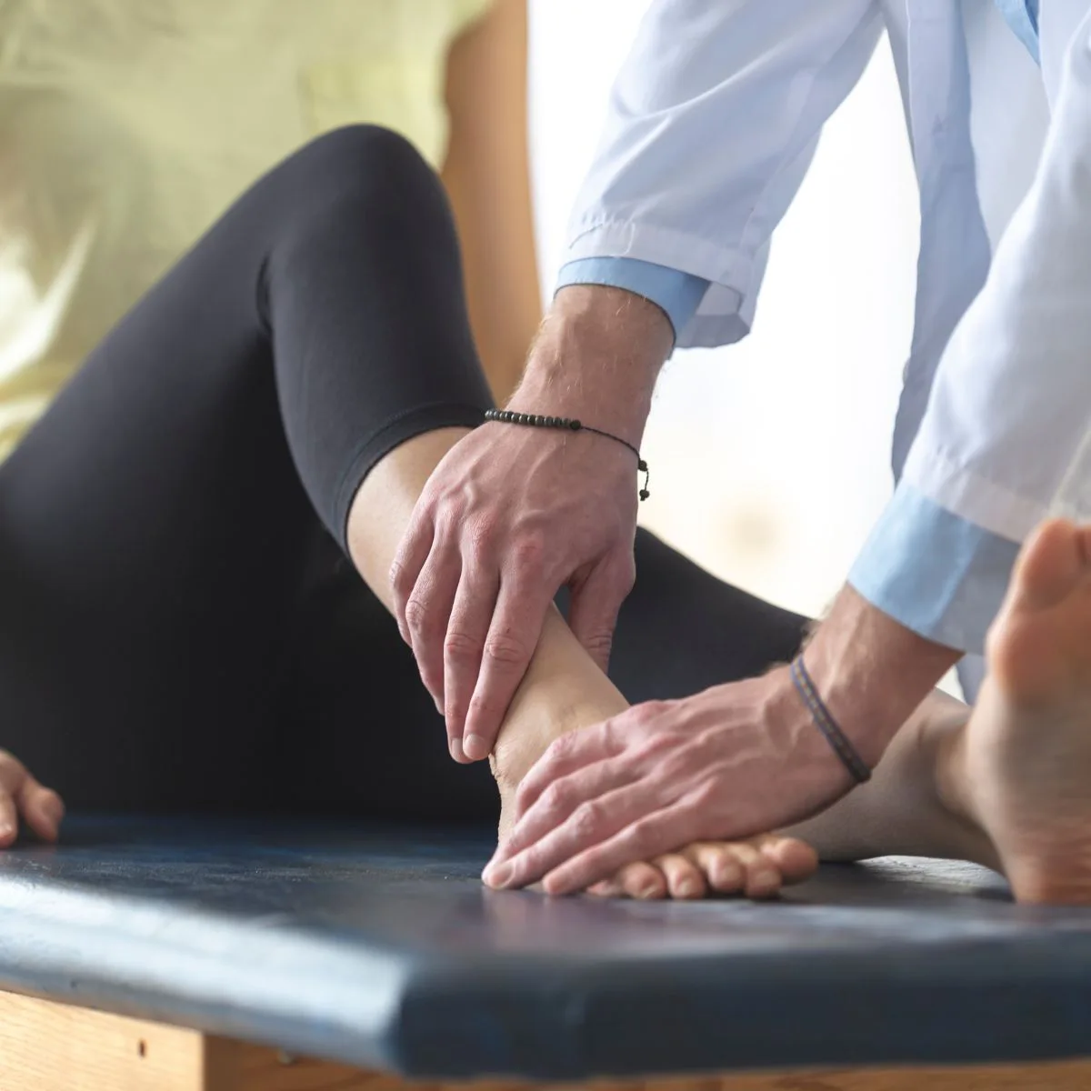 A healthcare professional examines a persons ankle while they sit on an examination table, focusing on the joint and applying gentle pressure with both hands.