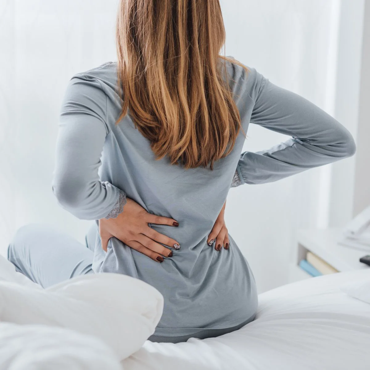 A woman with long hair sits on a bed facing away, wearing light blue pajamas, and holds her lower back with both hands, suggesting discomfort or back pain.