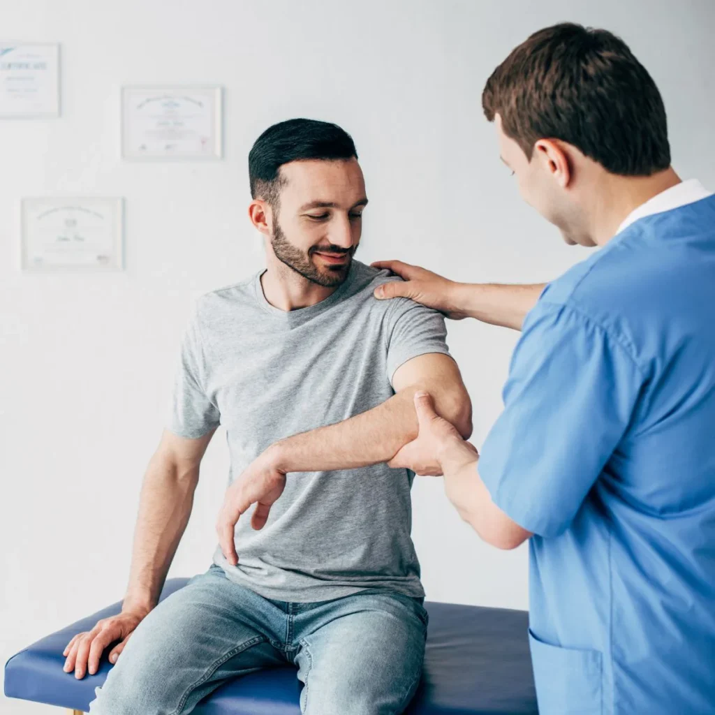 A healthcare professional in blue scrubs examines a mans arm while he sits on an exam table. The man is wearing a gray t-shirt and jeans and looks at his arm during the examination in a medical office.