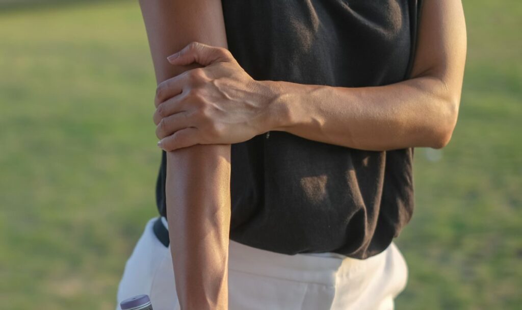 A person wearing a black sleeveless top and white pants holds their left forearm with their right hand, standing outdoors on a grassy area.