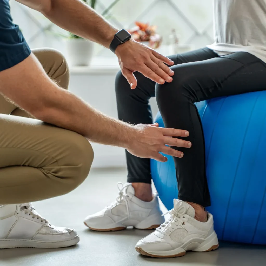 A person sits on a blue exercise ball while a physical therapist in beige pants and white sneakers assesses their knee. The therapist gently places their hands near the person’s leg.