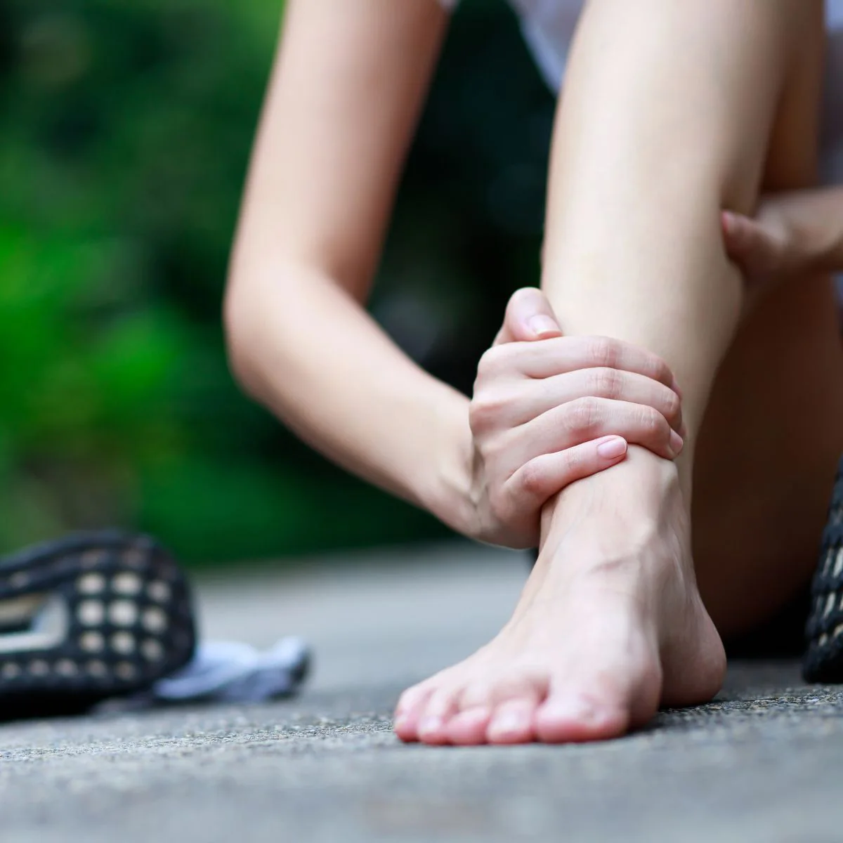 A person sitting on the ground outdoors holds their ankle with both hands, appearing to be in pain. One shoe is off, and the background is blurred greenery.