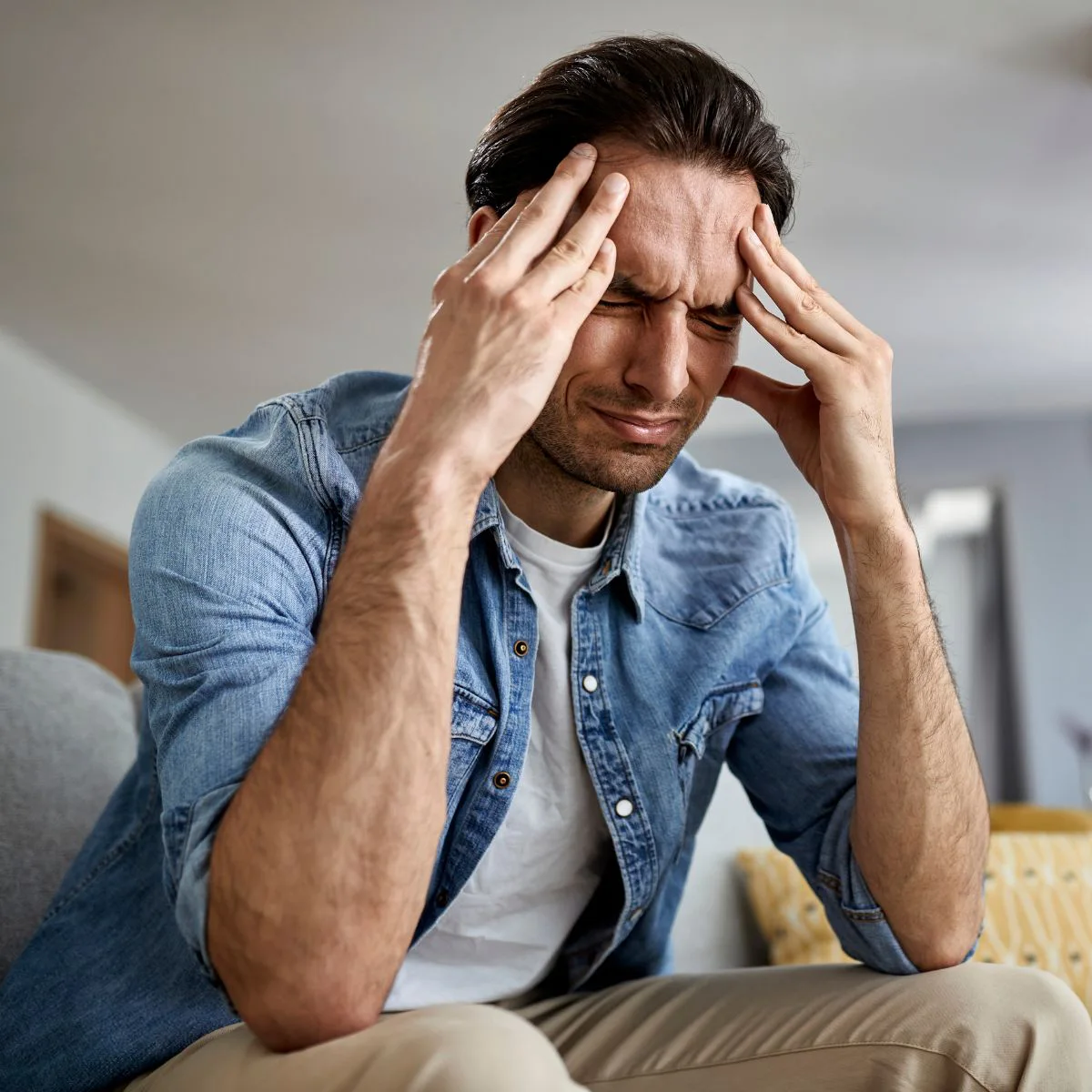 A man sitting on a couch with his eyes closed, pressing his temples with his fingers and appearing to be in pain or stressed. He is wearing a denim shirt and beige pants.