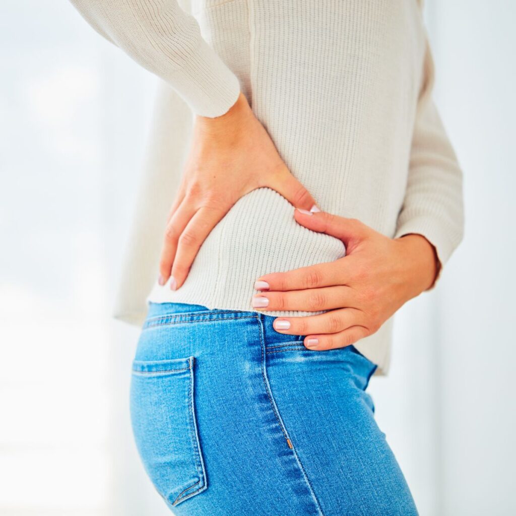A person wearing a light sweater and blue jeans holds their hip with both hands, appearing to be in discomfort or pain.