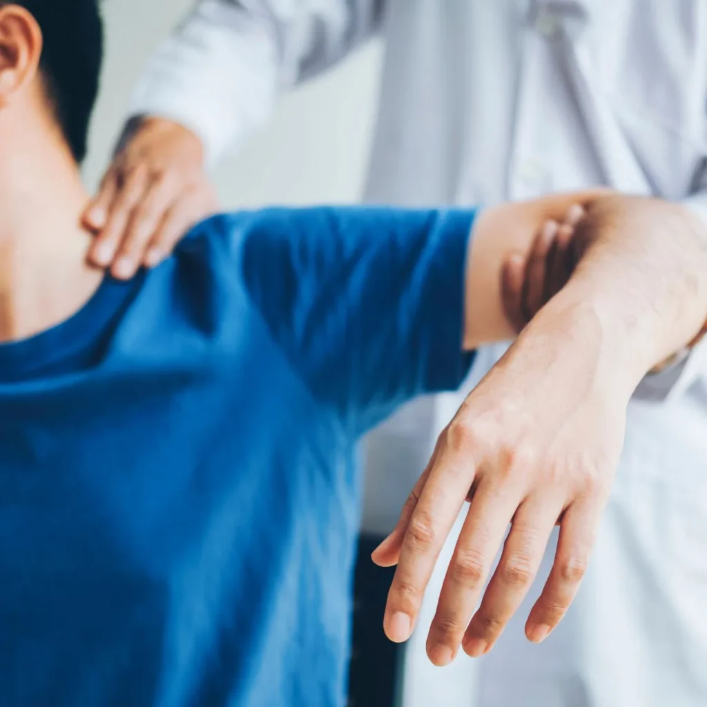 A person in a blue shirt receives a shoulder and arm examination or therapy from a healthcare professional wearing a white coat. The professional is gently manipulating the patients arm and shoulder.