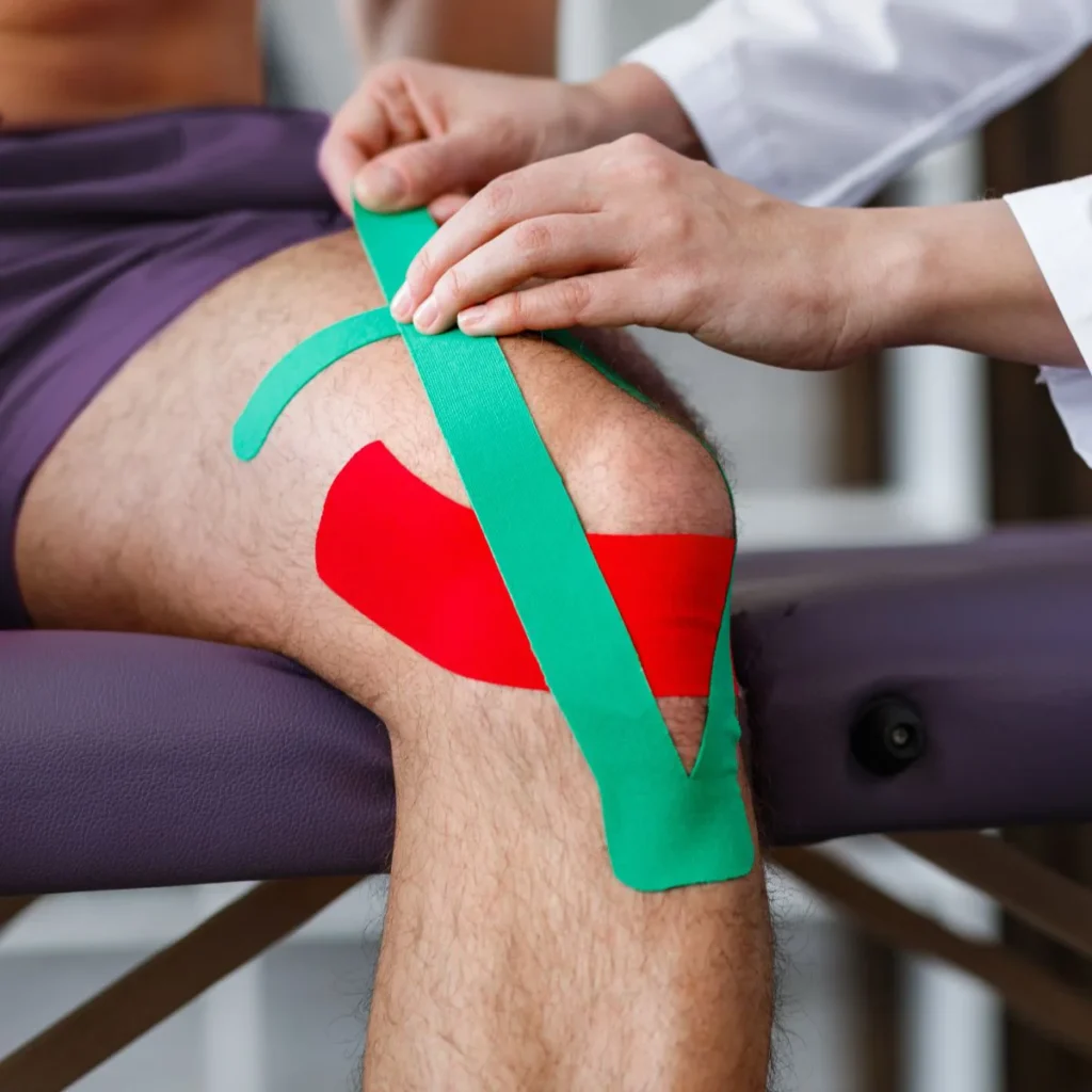 A person’s knee is being taped with colorful kinesiology tape—one green strip and one red—by a healthcare professional, likely for support or injury recovery. The person is seated on an examination table.