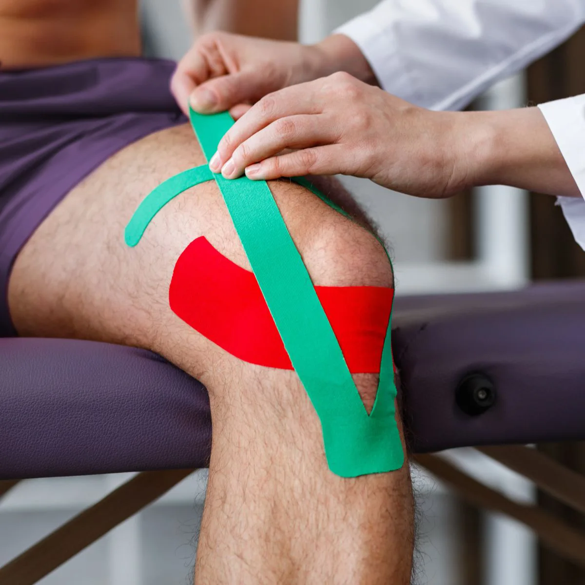 A person’s knee is being taped with colorful kinesiology tape—one green strip and one red—by a healthcare professional, likely for support or injury recovery. The person is seated on an examination table.