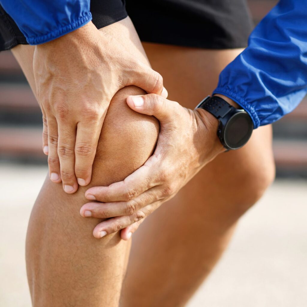 A person wearing a blue jacket and black shorts is holding their knee with both hands, appearing to experience knee pain or discomfort outdoors.
