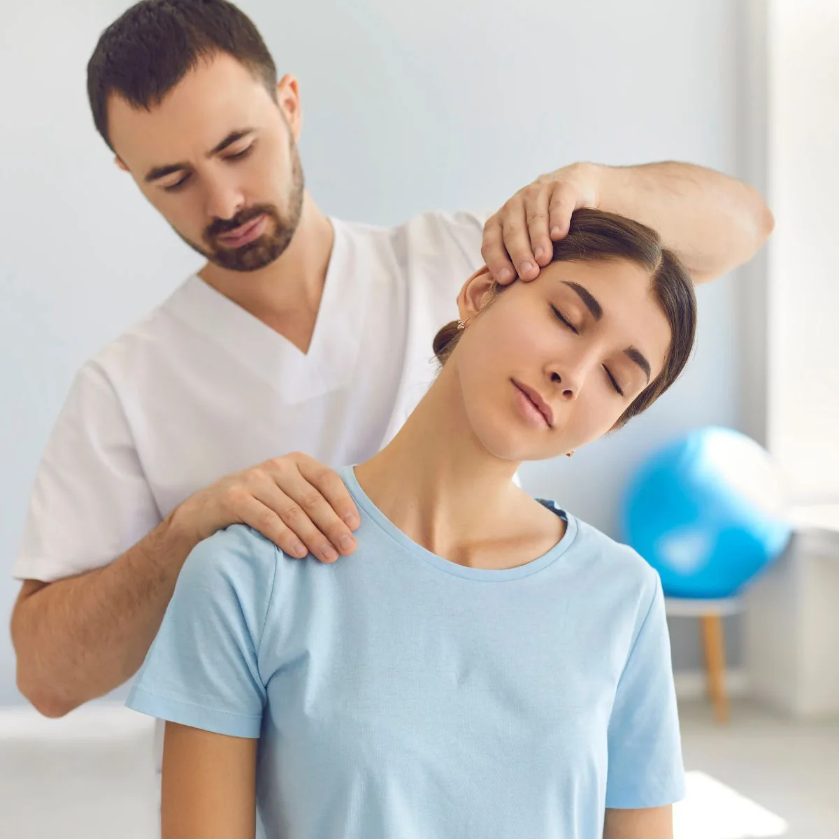 A physical therapist gently stretches a woman’s neck while she sits with her eyes closed, wearing a light blue shirt, in a bright therapy room with exercise equipment in the background.