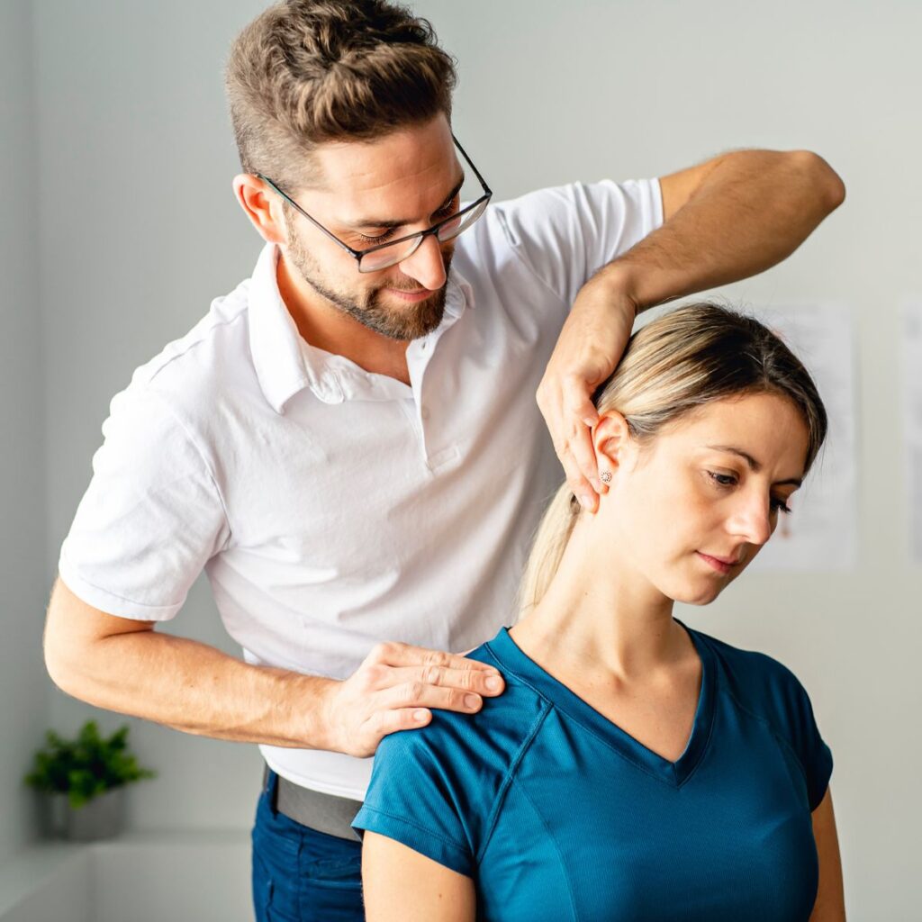 A man in glasses and a white shirt assists a woman in a blue shirt with a neck stretch, gently holding her head and shoulder in a bright, clinical setting.