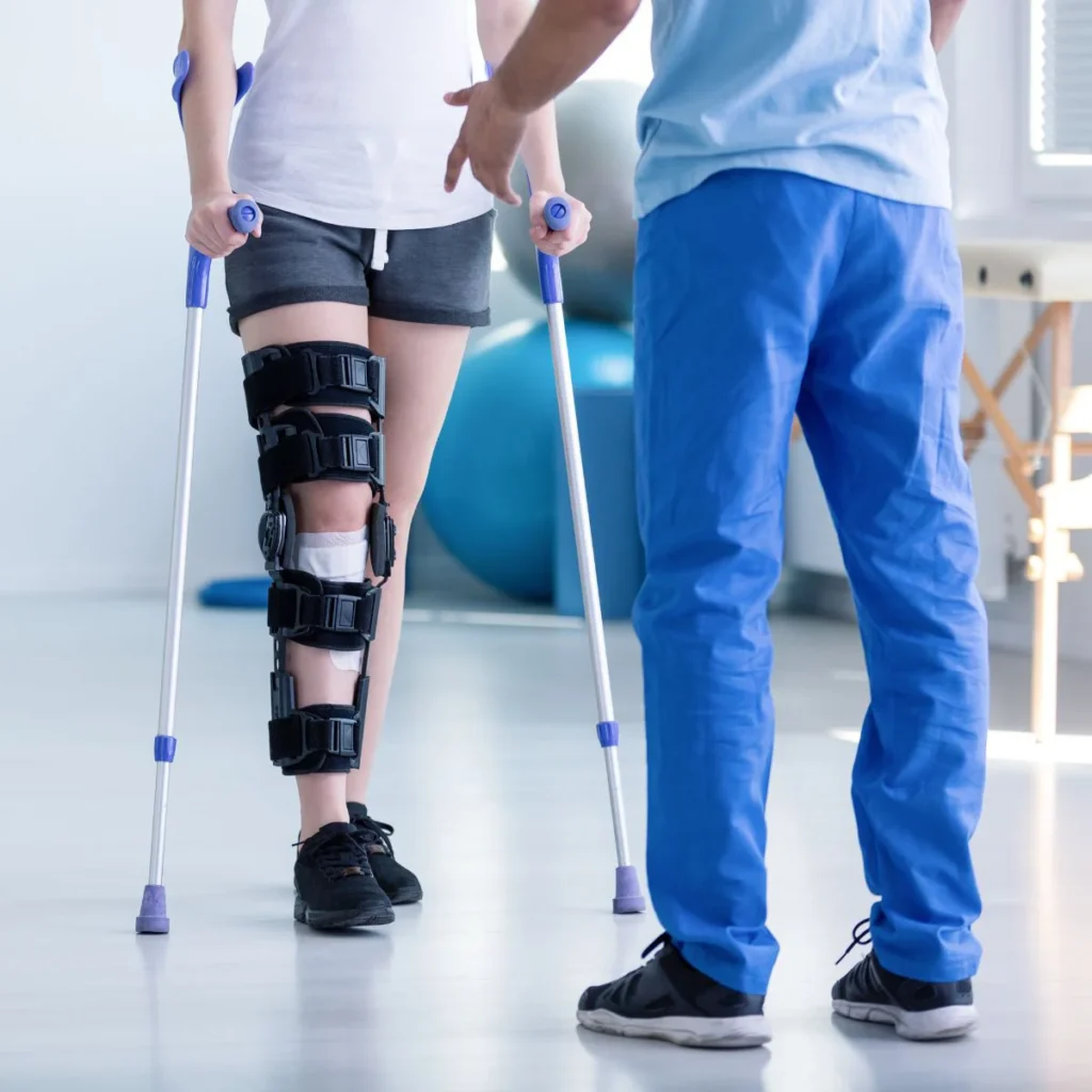 A person with a knee brace and crutches stands while a healthcare professional in blue scrubs offers assistance in a medical or rehabilitation setting.