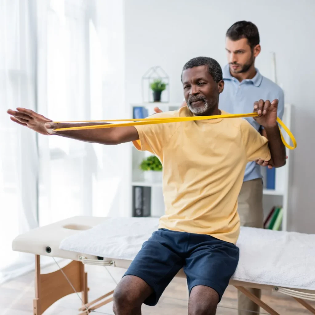 An older man in a yellow shirt uses a resistance band for arm exercises while seated on a therapy table, assisted by a physical therapist in a light-filled clinic.