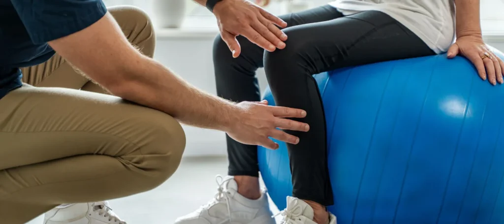 A person sits on a large blue exercise ball while another person, possibly a physical therapist, supports and guides their leg during a therapy or exercise session.