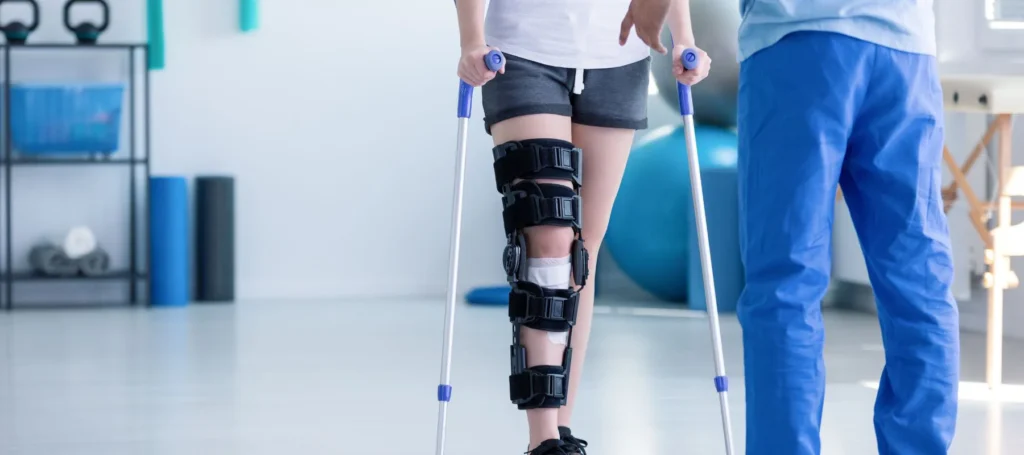 A person with a knee brace on their right leg uses crutches while a healthcare professional stands nearby in a bright rehabilitation clinic.