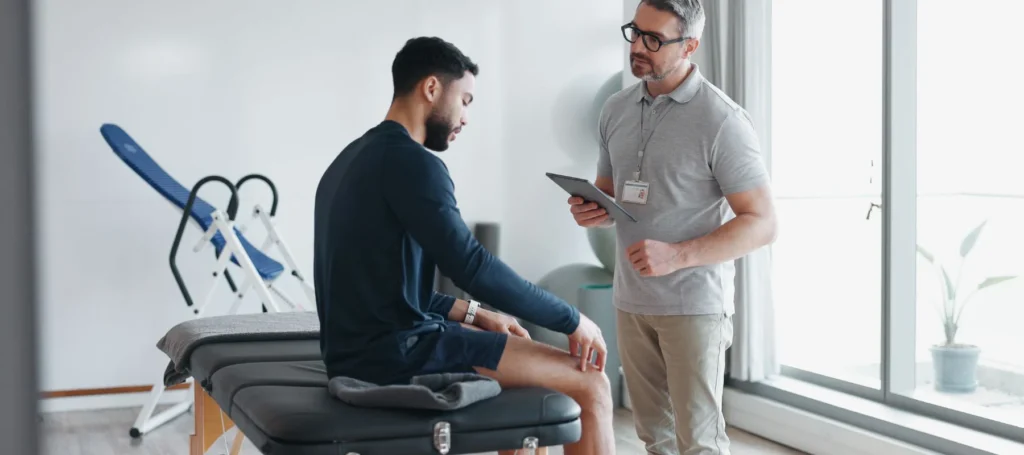 A man in athletic wear sits on an exam table, holding his knee, while a healthcare professional with a tablet talks to him in a bright, modern clinic.