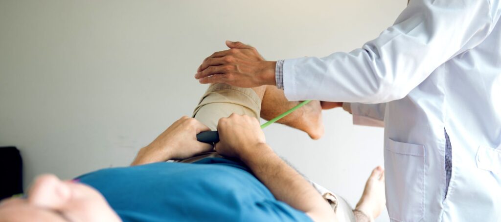 A doctor in a white coat examines the knee of a patient lying down. The patients knee is wrapped in bandages and the doctor appears to be measuring or assessing the injury.