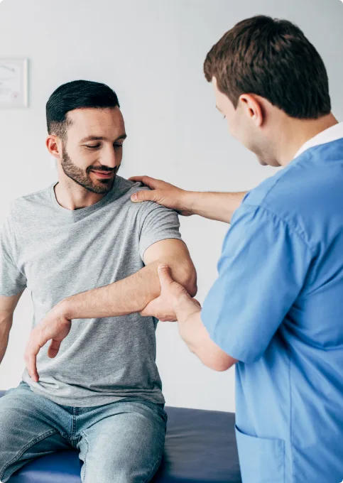 A healthcare professional in blue scrubs examines a seated mans arm, gently holding and moving his elbow while the man looks at his arm and smiles. The setting appears to be a medical office.