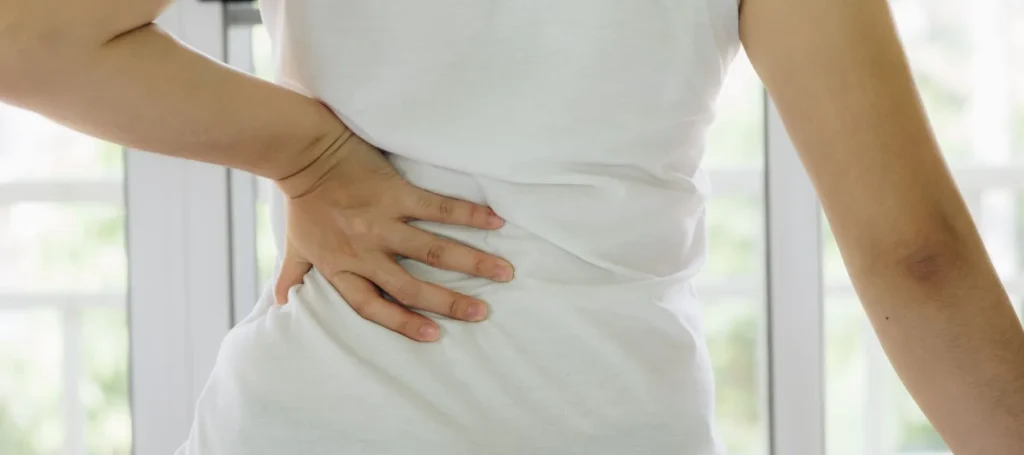 A person in a white shirt stands indoors with one hand placed on their lower back, appearing to experience back pain or discomfort.