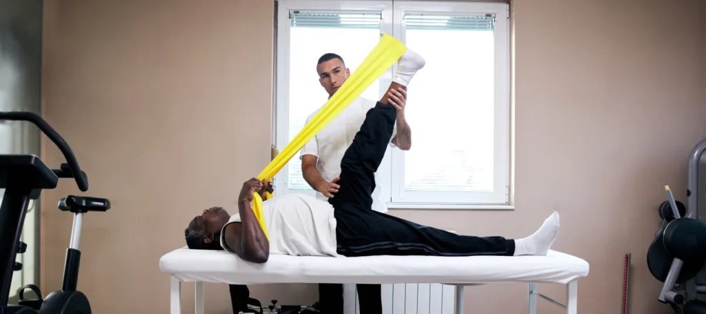 A physical therapist helps a man lying on a table stretch his leg using a yellow resistance band in a well-lit room with exercise equipment.
