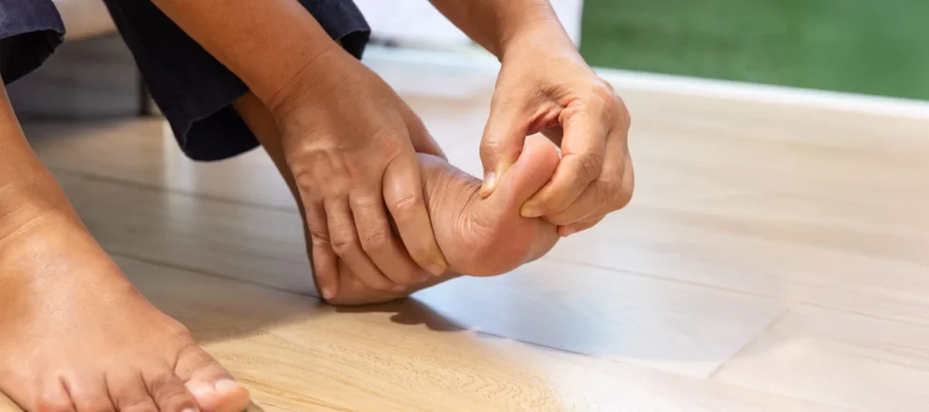 A person sitting on the floor is holding and pressing the toes of their right foot with both hands, possibly indicating discomfort or pain. The floor is wooden and there is a blurred green background.
