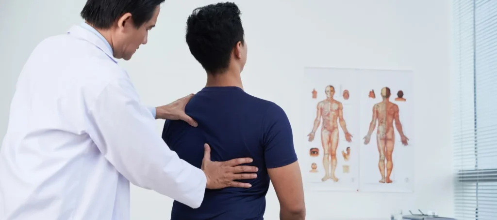 A doctor examines a patients back in a medical office. The patient is sitting, facing away. Anatomical charts are visible on the wall in the background.
