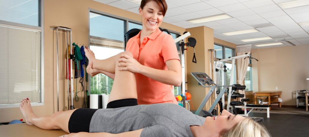 A physical therapist helps a smiling woman lying on a table perform a leg stretch in a bright, well-equipped rehabilitation clinic.